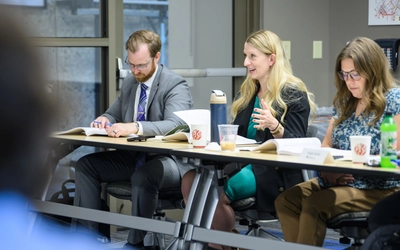 A blonde woman in a green dress and blazer gestures while speaking during a group meeting, seated between two colleagues reviewing materials at a conference table.