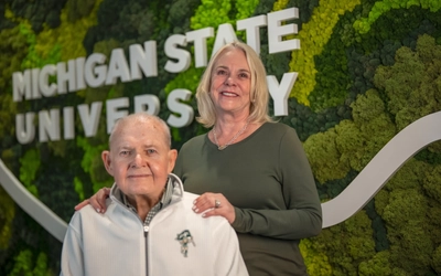Two adults pose for a photo in front on a Michigan State University backdrop with the MSU writing and greenery.