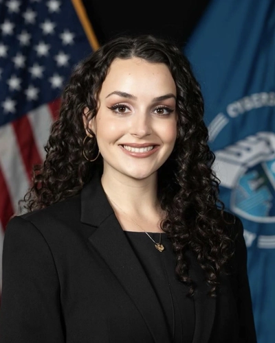 Professional headshot of Rina Rakipi in which she smiles with long, dark curly hair wearing a black blazer, standing in front of the American flag and a blue agency flag.