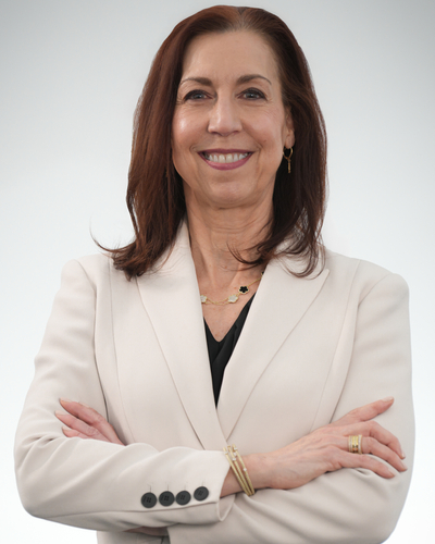 Person stands with arms crossed, wearing a light-colored blazer over a dark top, photographed against a plain light background.
