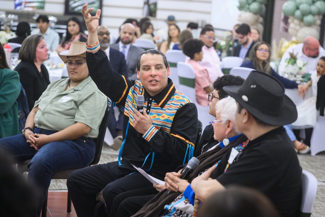 people sit in a circle, one man has his arm raised and is talking into a mic as people listen