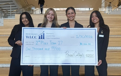 Vivian Tieu, Megan Howarth, Sofia Poulos and Anvi Thakur stand at the bottom of a stair case holding a large check.