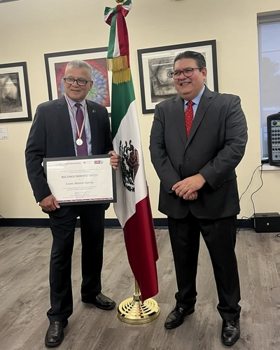 Louis Alonzo Garcia displays his Ohtli Award certificate next to an official at a presentation event next to a Mexican flag.