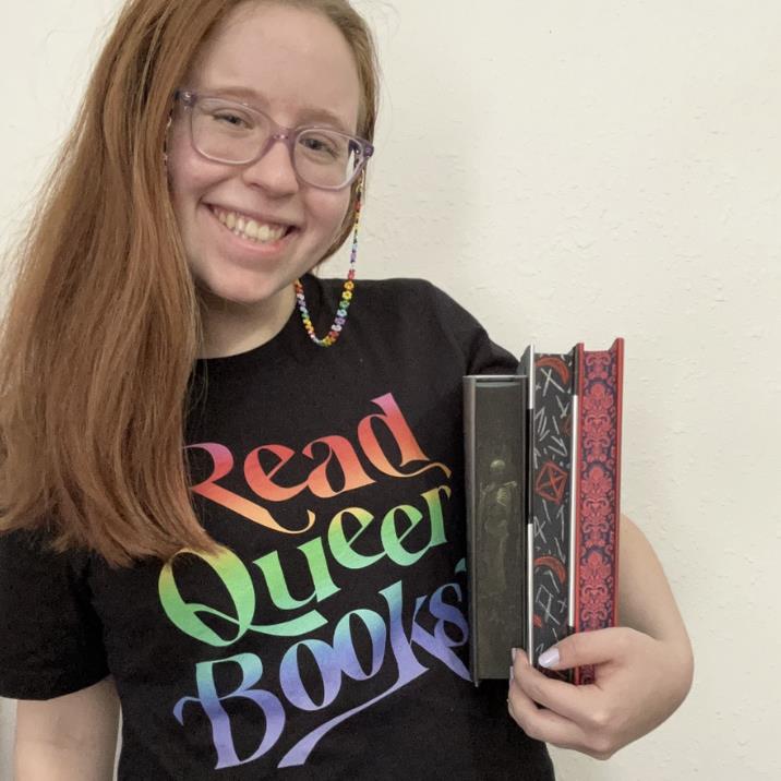 person smiling and holding book items wearing a read, queer books shirt