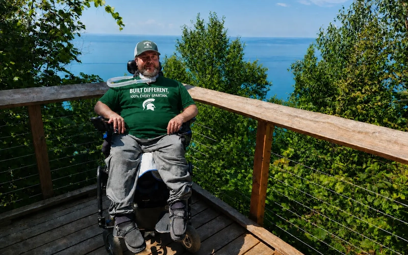 Mo Gerhardt in a wheelchair wearing a green T-shirt with "Built Different: 100% Every Spartan" on the front while on a wooden overlook by a large blue lake.