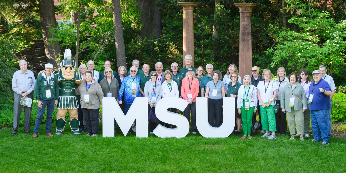 Outside in a garden, a large group of people stand behind large, white letters for MSU.