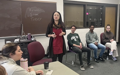 An image of Jia Bin standing at the front of a classroom with students sitting before her, leading the JRN 492 course stuttering panel.