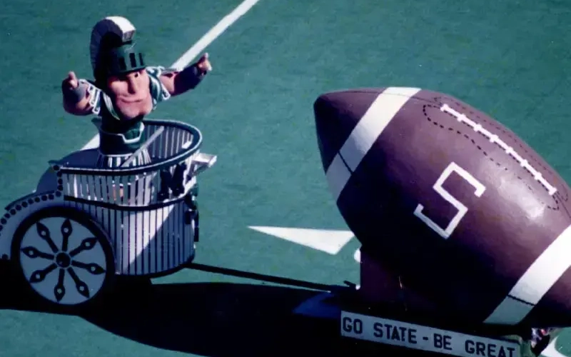 New Sparty mascot debuting on football feild at Spartan stadium in 1989. The mascot is in a chariot attached to a paper mache football, on a platform that says "Go State, be great"