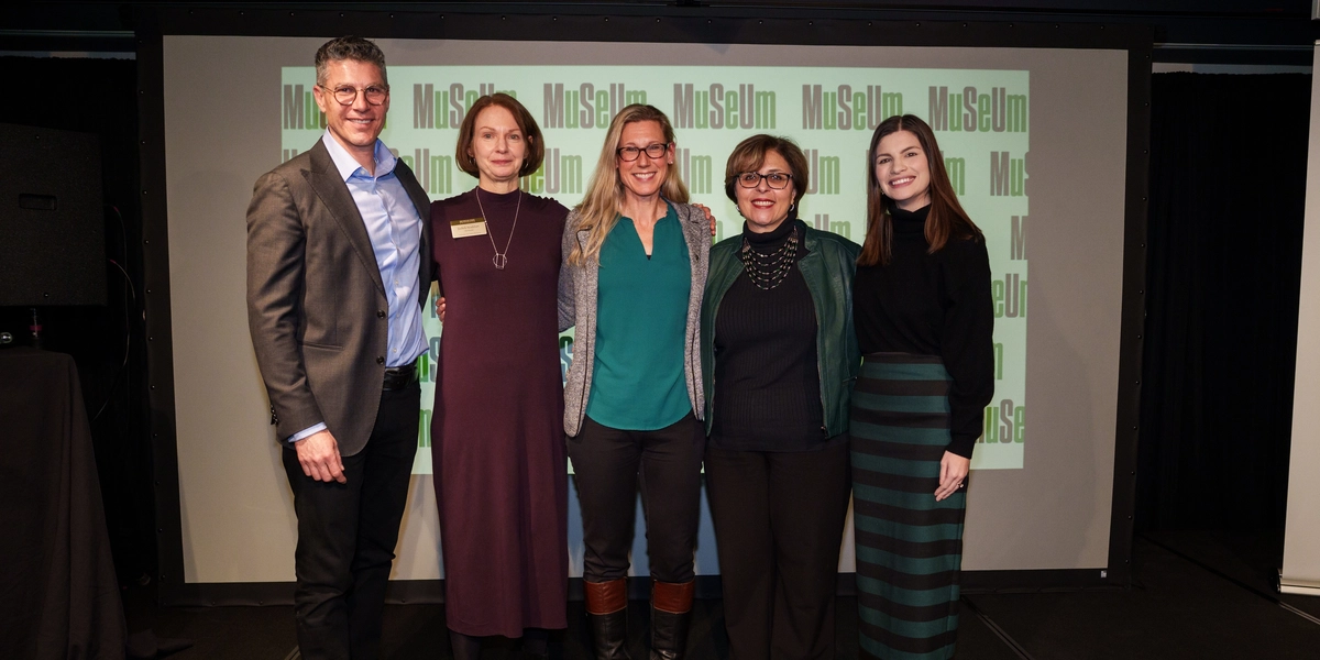 A group of five people stands together on a stage in front of a screen displaying a repeated “Museum” logo. They are posed side by side, dressed in business or business‑casual attire, and smiling for the photo. The background is a dimly lit event space with black curtains and a projector screen behind them.