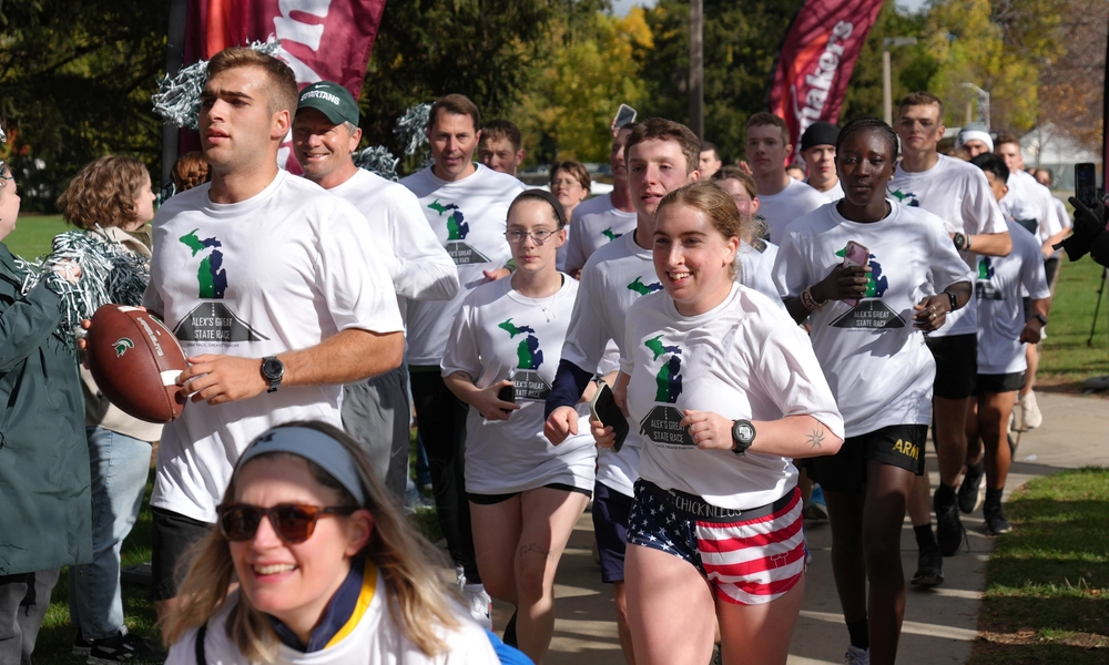 A group of runners and a hand cyclist wearing white t-shirts for Alex's Great State Race move toward the finish line with onlookers cheering them on.