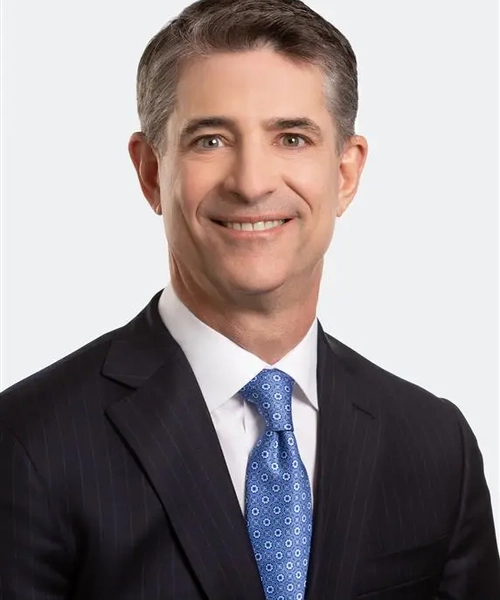 A man wearing a dark blue pinstripe suit with a blue patterned tie and a white button down shirt against a white background.