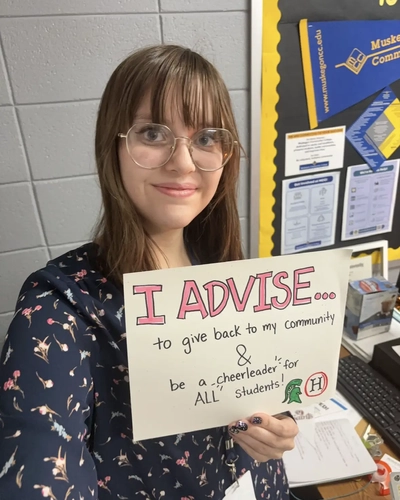 A young woman with brown hair, bangs, and round glasses smiling while holding a hand-drawn sign. The sign reads, "I ADVISE... to give back to my community & be a 'cheerleader' for ALL students!" It features small logos for Michigan State University and a high school. She is standing in front of a school bulletin board with college pennants.