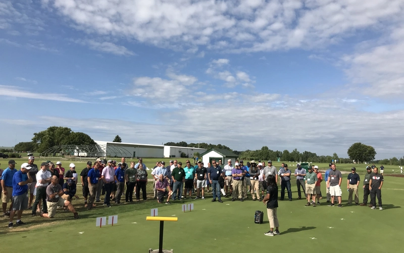 Presenter speaks to an outdoor crowd gathered on a turfgrass field.