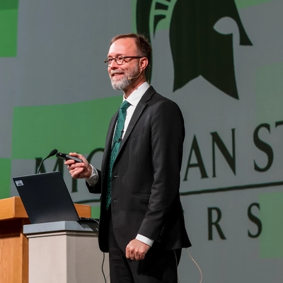 A speaker in a suit and green tie standing at a podium with a laptop during a presentation.