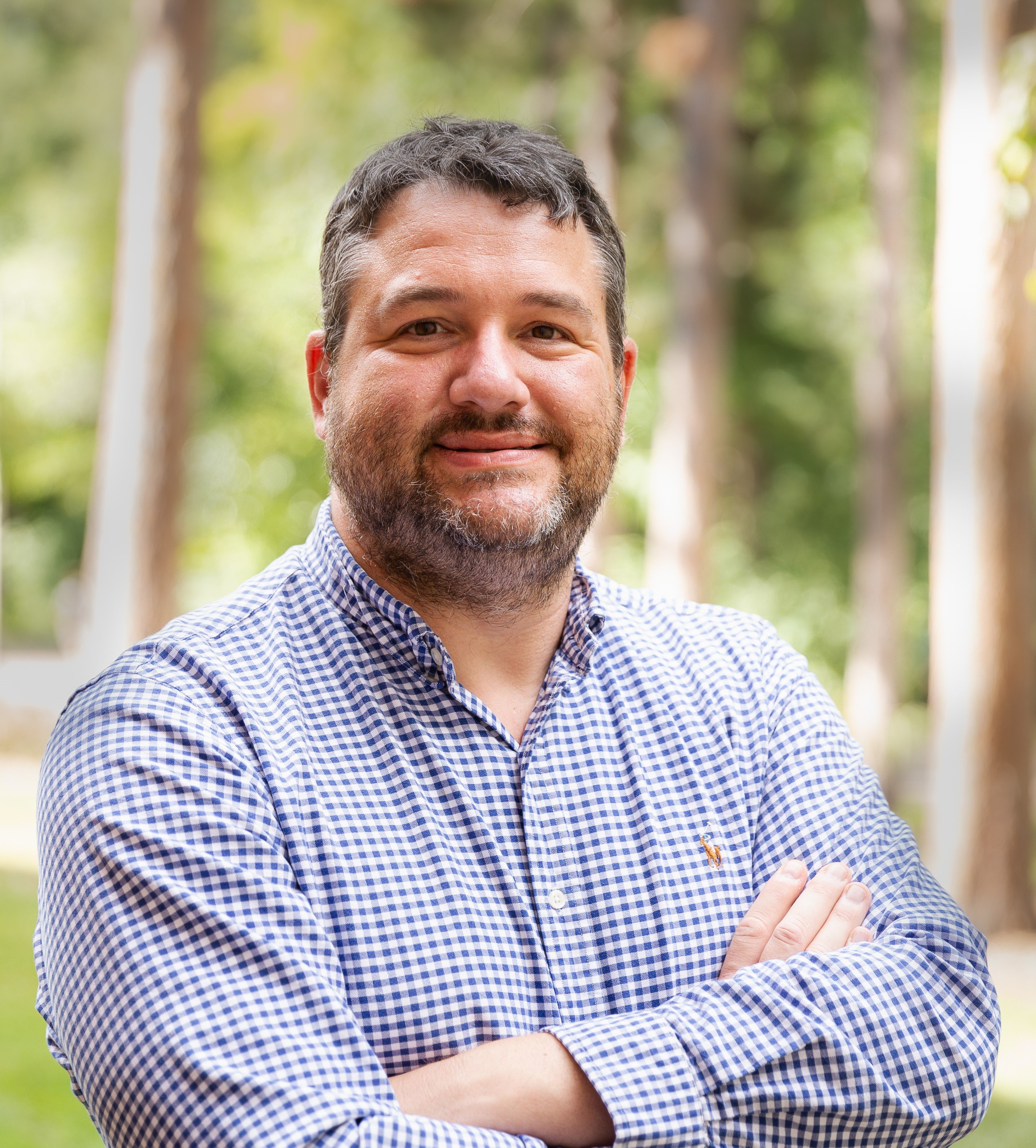 Smiling man with short dark hair and a beard, arms crossed, wearing a blue checkered shirt outdoors.