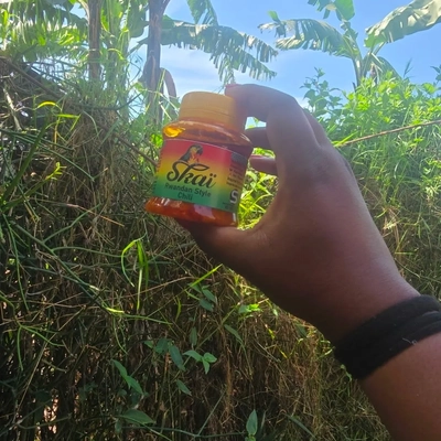A hand holds a bottle of Skai Rwandan-style chili sauce in front of green vegetation and banana trees in Rwanda.