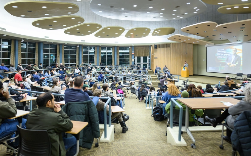 Attendees sit in curved, tiered rows in Erickson Hall, facing a speaker at a lectern beside a large projection screen.