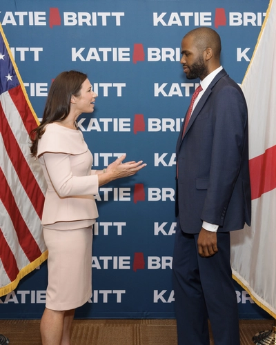 A man in a suit speaks with Senator Katie Britt in front of a backdrop with her name and two flags.