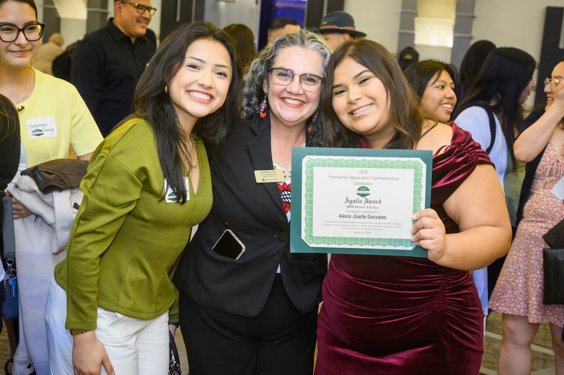 Three women pose or a photo in a crowded room, holding a certificate