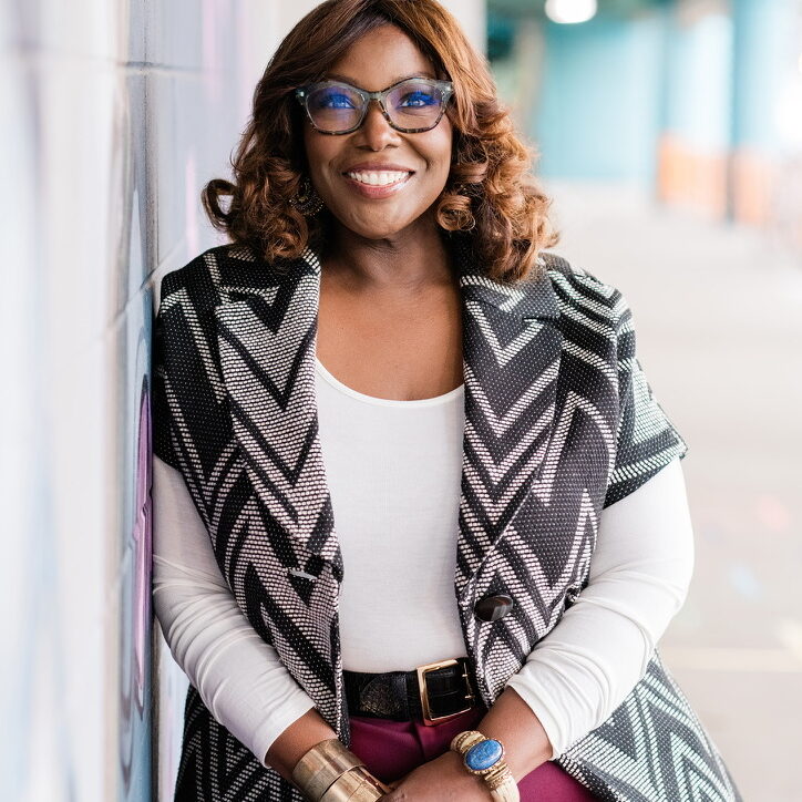 Kinitra Brooks wearing glasses and a black-and-white patterned vest, leaning against a wall in a bright hallway.