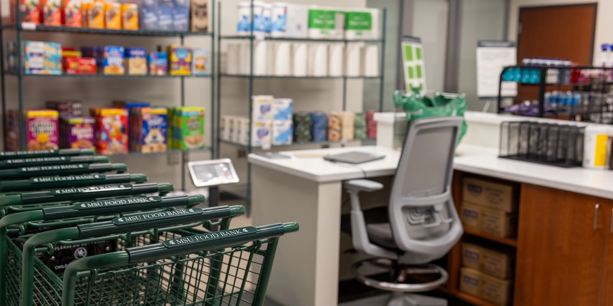 A photo of the interior of Spartan Shelf. Green carts in the foreground, a desk and grocery shelves lined with products in the background.