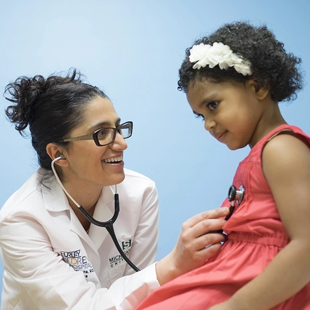 A pediatrician smiles while listening to a young girl's heartbeat with a stethoscope during a checkup.