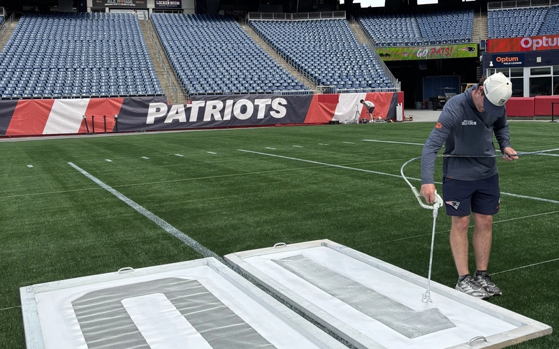 A field crew member sprays white paint over large number stencils on the turf, with a Patriots banner behind him.