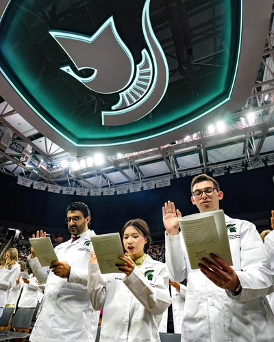 A celebratory moment during the Michigan State University College of Osteopathic Medicine White Coat Ceremony, captured on stage at the Breslin Center. A smiling medical student in a suit and tie, holding a program and raising their right hand to take an oath, stands between two other smiling medical students in white coats. The background features warm stage lighting and acoustic wood paneling in an auditorium setting.