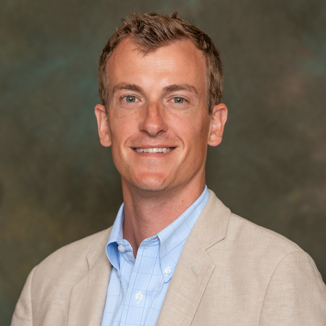 Person wearing a light beige blazer over a light blue button-up shirt with a subtle checkered pattern, posed against a mottled brown and green studio backdrop.