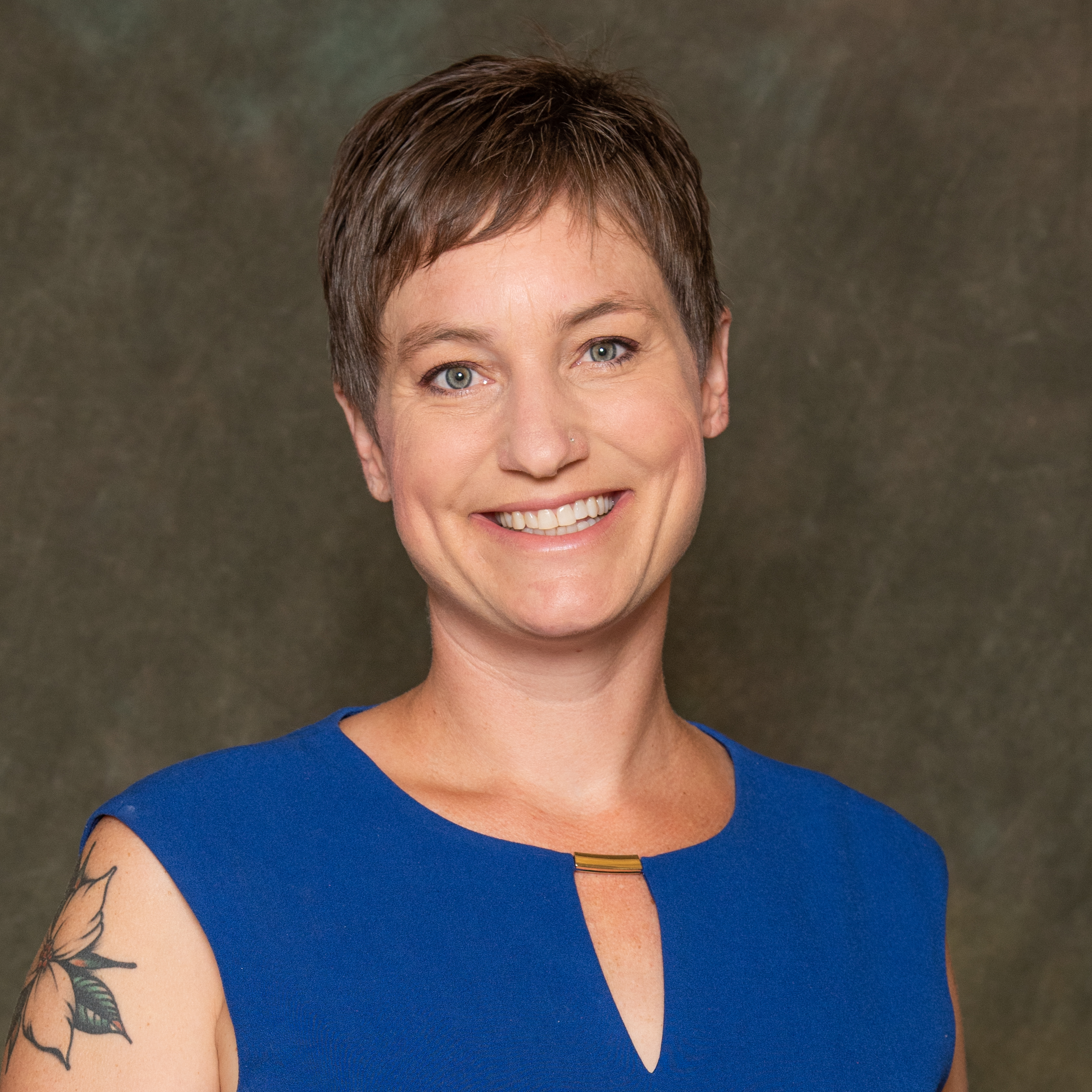 Person wearing a sleeveless royal blue top with a keyhole neckline featuring a small gold bar detail, posed against a mottled dark green and brown studio backdrop. A floral tattoo is visible on the upper left arm.
