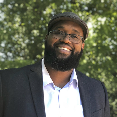 Glenn Chambers wearing a dark suit jacket over a light-colored dress shirt. He has glasses and is also wearing a flat cap hat. He is standing outdoors with green foliage in the background.