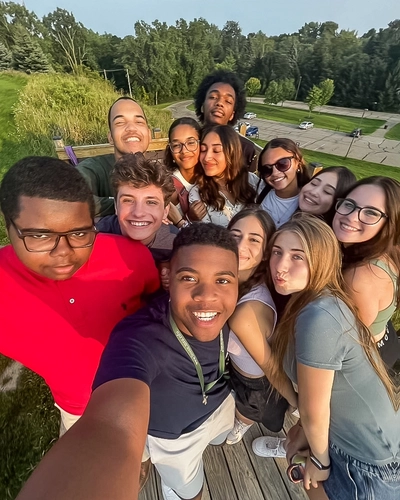 A group of students poses in a selfie on top of a wooden platform on a hill with a parking lot in the background below them