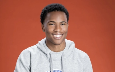 A smiling young adult in a New York Knicks hooded sweatshirt posing with a red background