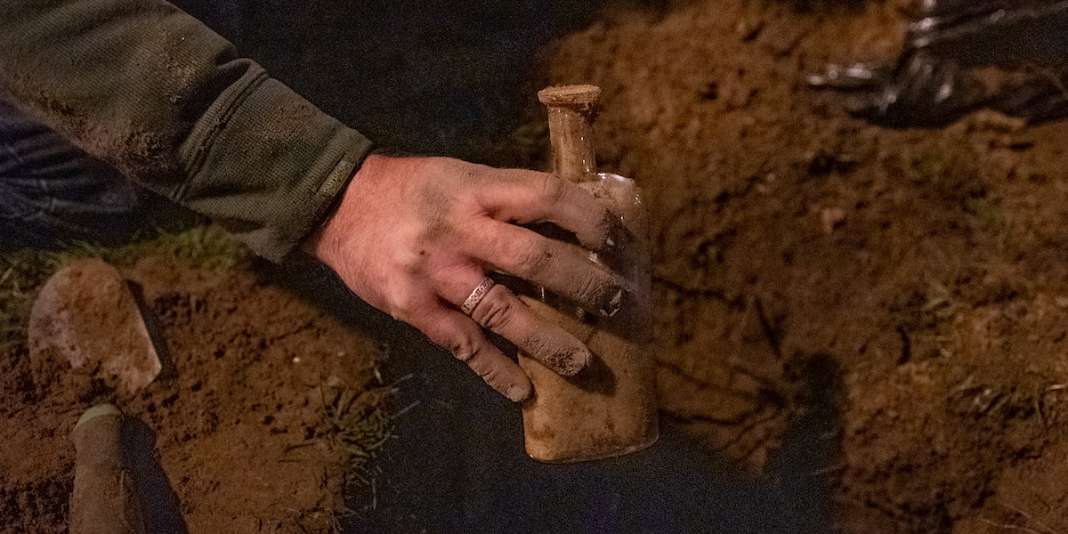 A hand pulls a glass bottle covered with dirt from a hole