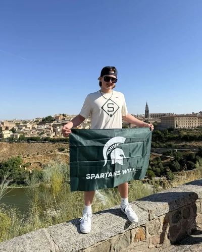 A person stands on a stone ledge holding a green flag with the Michigan State University Spartan helmet logo and the words "SPARTANS WILL." The person is wearing a white t-shirt with an "S" logo, white shorts, and white sneakers. The background shows a scenic view of an old city with historic buildings and a river.
