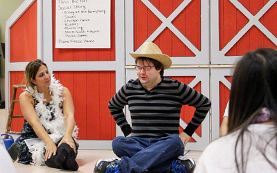 A group of adults sits on the floor in front of a red barn-themed backdrop. One man wearing a striped shirt and straw hat is speaking, while a woman beside him, dressed in a feather boa, listens.