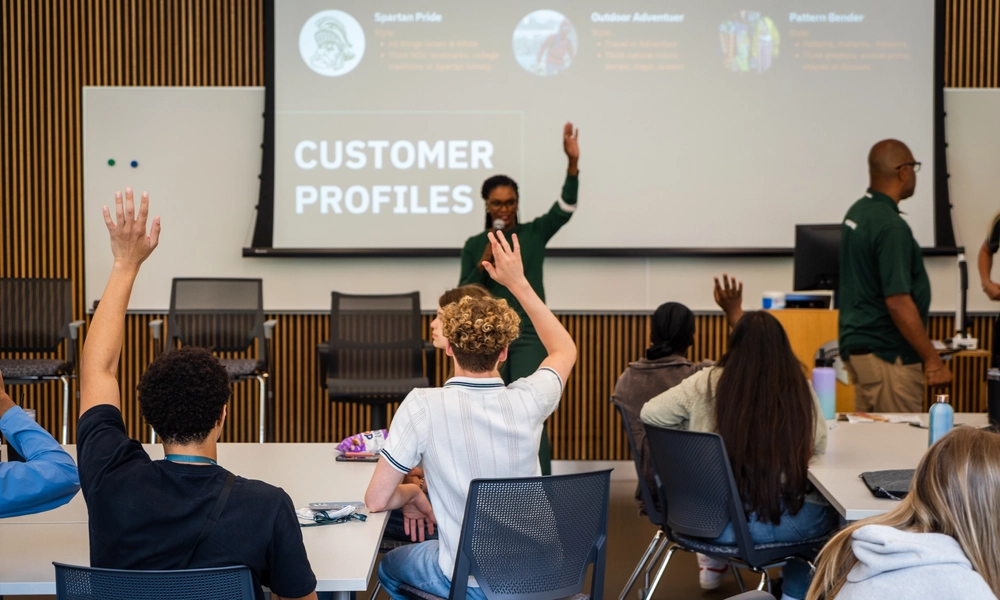 Classroom setting with several people seated at tables facing a large screen displaying a presentation titled ‘Customer Profiles.’ Some participants have their hands raised, and two presenters stand near the front of the room