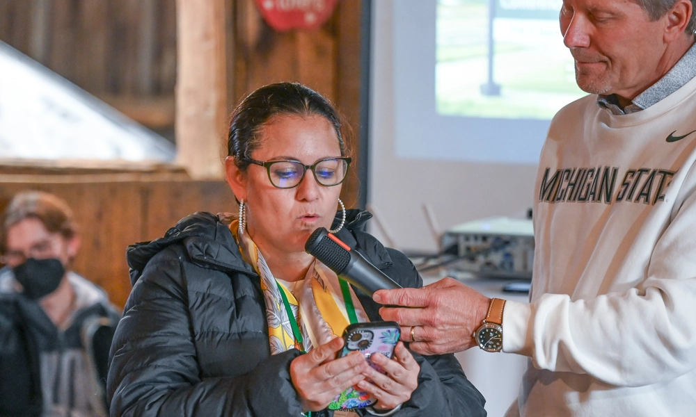 A person holds a smartphone while speaking into a microphone during an indoor event. Another individual is extending the microphone toward the speaker. The setting includes wooden walls, a projector displaying an image on a screen, and a table with equipment in the background. The person holding the microphone is wearing a sweatshirt with the words “Michigan State” visible. Several people are seated in the background.