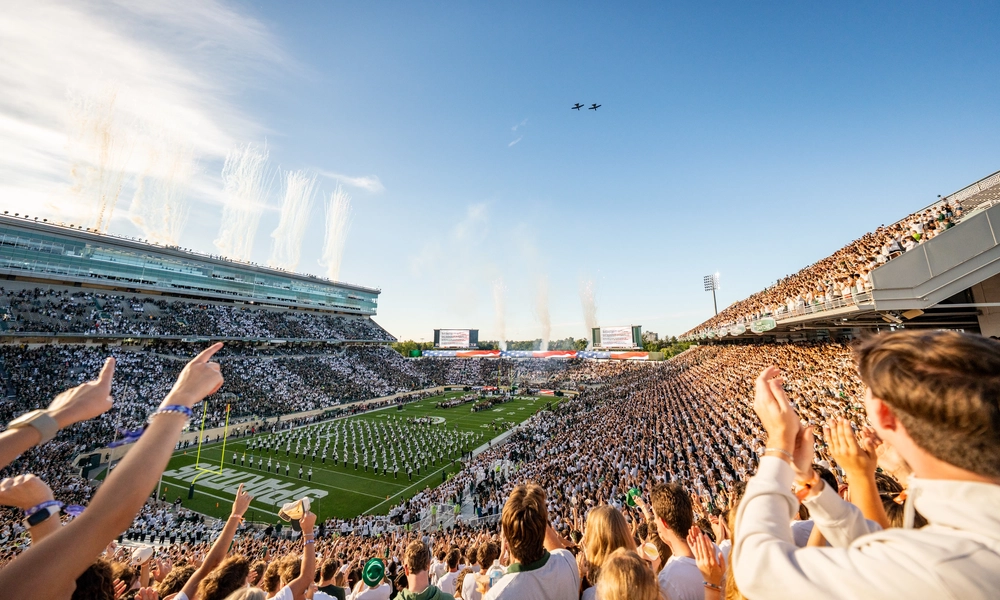 Two jets fly over a full Spartan Stadium on a sunny day as plumes of smoke shoot up from the stadium.