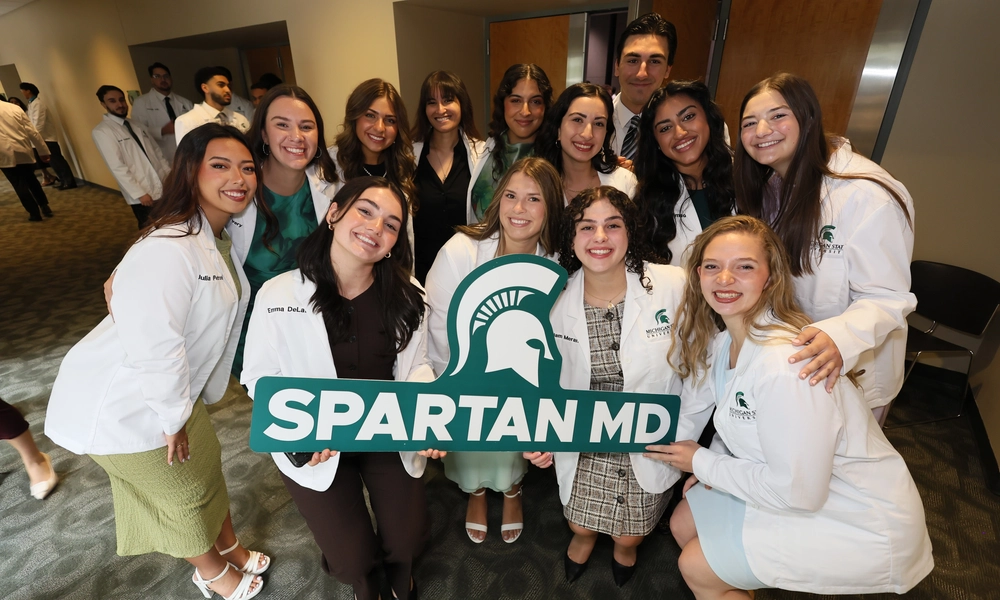 A group of Michigan State University medical students in white coats smile and pose together holding a large green “Spartan MD” sign during the White Coat Ceremony.