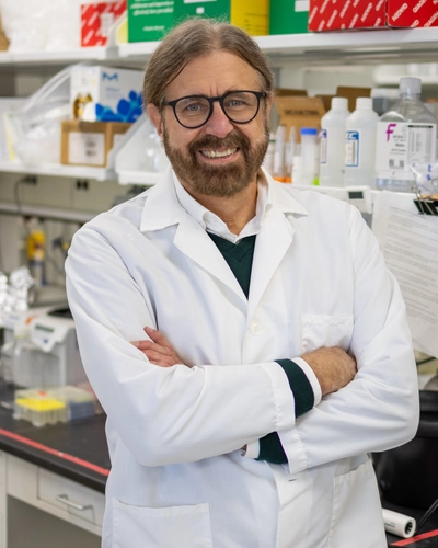 A person wearing a white lab coat stands in a laboratory with arms crossed. The background shows various laboratory equipment, including bottles, boxes, and shelves filled with supplies.
