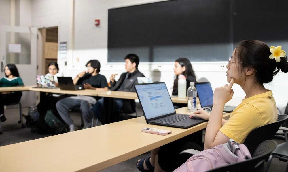 A group of students sit at desks discussing topics with computers and phones in front of them