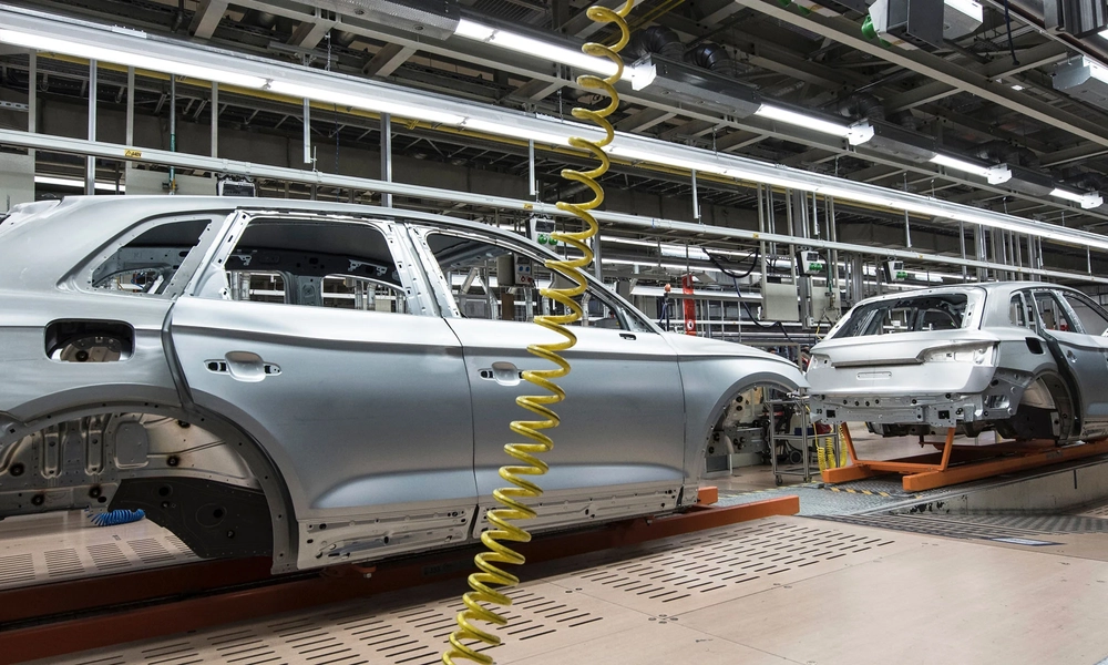 A car manufacturing assembly line with several partially assembled silver car bodies on conveyor belts. The factory has overhead lighting, yellow coiled air hoses hanging from the ceiling, and various machinery and equipment used in the assembly process.
