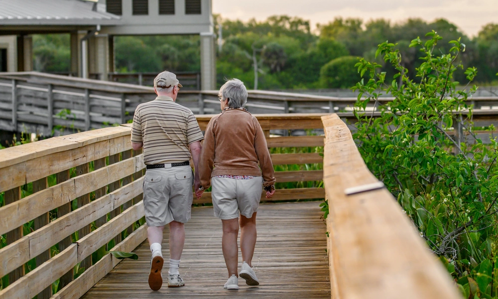 An elderly couple walking hand in hand on a wooden boardwalk surrounded by greenery, with a building featuring a metal roof and louvered windows in the background.