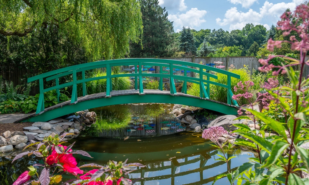 A small green arched bridge spans a tranquil pond with lush greenery and flowers surrounding it at the Michigan 4-H Children