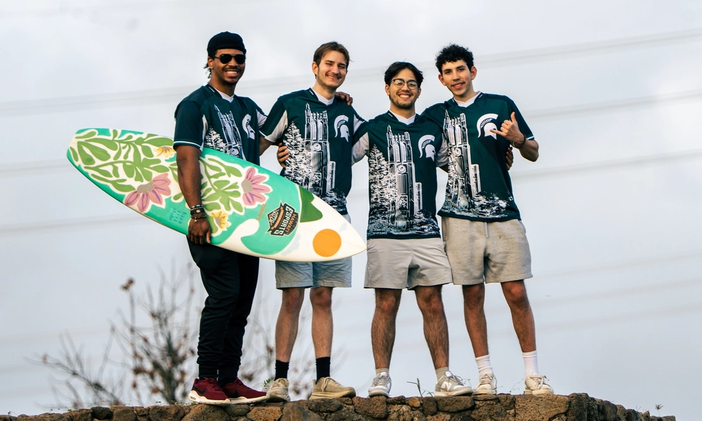 Four MSU esports players pose. The player on the left is holding a surfboard, the trophy for winning a tournament.