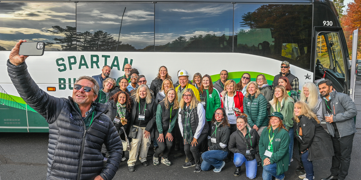 A large group of people pose for a photo in front of a white and green tour bus labeled “Spartan Bus Tour.” One person in the foreground holds up a smartphone to take a selfie with the group. The bus is parked on an asphalt lot surrounded by trees with autumn foliage, and a colorful wood figure is visible behind of the bus.