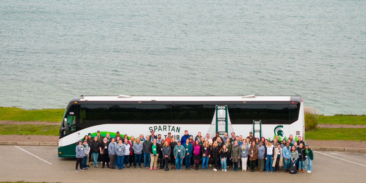 A large group of people stands in front of a white and green tour bus labeled “Spartan Bus Tour,” parked near a waterfront. Behind them, the Mackinac Bridge stretches across the water under a cloudy sky. The scene includes a paved parking area in the foreground and grassy shoreline leading to the water