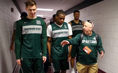 Michigan State University basketball players walk down a hallway with staff member Mex Carey, who is speaking animatedly and smiling.