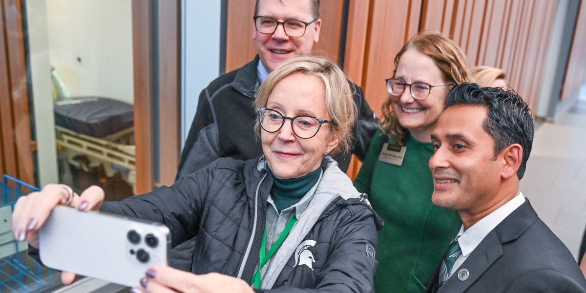 A group of people stand near a wall with vertical wooden panels, posing for a selfie. One person in the foreground holds a smartphone at arm’s length to capture the photo. Visible behind the group is a large window with a view of a room containing a bed or examination table.